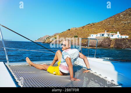 Fröhlicher junger Mann fühlt sich in den Sommerferien auf der Insel glücklich über den luxuriösen Segelbootjacht-Katamaran im türkisfarbenen Meer. Kaukasisches männliches Modell. Stockfoto