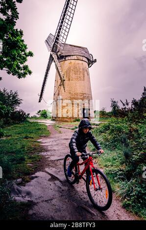 Der Junge, der mit dem roten Fahrrad im Regen unterwegs ist, passiert die alte historische Windmühle Bidston Hill Wirral UK Stockfoto