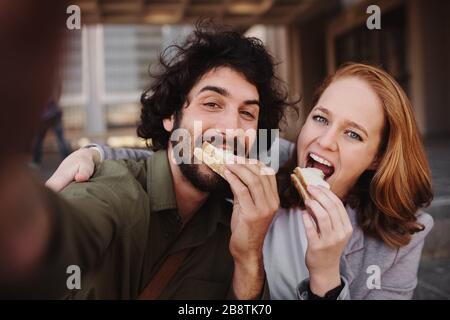 Fröhliches junges Berufspaar, das ein selfie mit dem Smartphone nimmt und dabei einen Bissen Sandwich packt Stockfoto