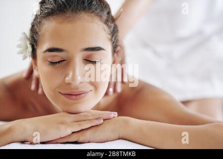 Hübsche junge Frau lächelt und schließt die Augen, wenn sie gerne wieder eine Massage im Spa-Salon bekommt Stockfoto