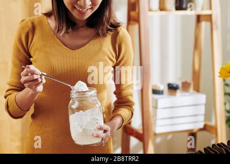 Lächelnde junge Frau, die bei der Herstellung duftender Seife zu Hause einen Löffel Sodalauge aus Glasbecher nimmt Stockfoto