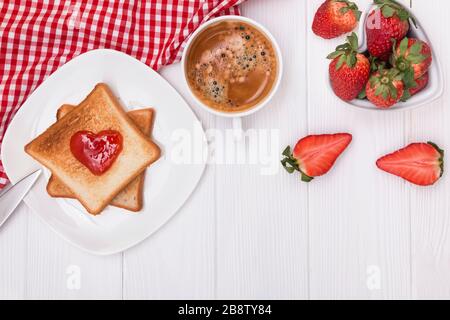 Brot und Marmelade in Herzform, Erdbeeren und Kaffee auf dem weißen Tisch Stockfoto