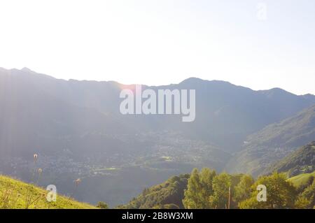 Ein kleines Land an der Bergwand mit der Sonne auf der Spitze, einer grünen Wiese und vielen Bäumen. Stockfoto