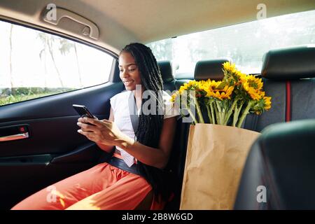 Schöne positive junge schwarze Frau, die nach dem Supermarkt im Taxi fährt und SMS von Freunden liest Stockfoto