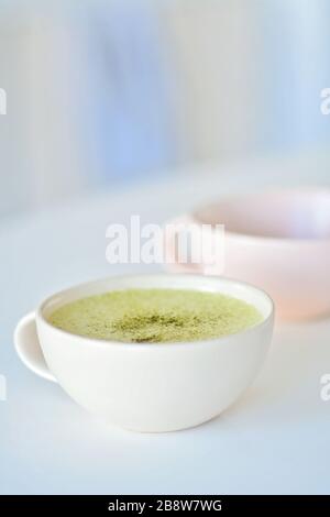 Weißer Becher mit Matchlatte auf weißem Tisch, einer hellrosa Tasse auf dem Hintergrund, Matcha-grüne Teelatte in einer Tasse. Draufsicht. Kopierbereich. Grüne Teematte Stockfoto