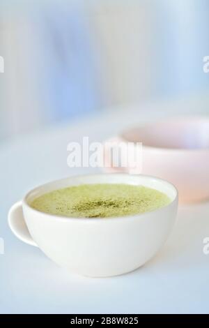 Weißer Becher mit Matchlatte auf weißem Tisch, einer hellrosa Tasse auf dem Hintergrund, Matcha-grüne Teelatte in einer Tasse. Draufsicht. Kopierbereich. Grüne Teematte Stockfoto