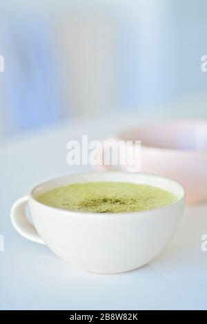 Weißer Becher mit Matchlatte auf weißem Tisch, einer hellrosa Tasse auf dem Hintergrund, Matcha-grüne Teelatte in einer Tasse. Draufsicht. Kopierbereich. Grüne Teematte Stockfoto