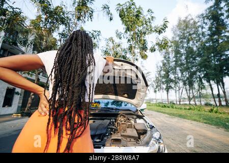 Junge Frau mit Dreadlocks, die die Motorhaube öffnet und versucht herauszufinden, was mit dem Auto passiert, Blick von hinten Stockfoto