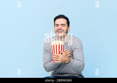 Junger Mann, der Popcorn lachen hält, während er einen Film sieht, der auf hellblauem Hintergrund isoliert ist Stockfoto