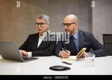 Portrait zweier erfolgreicher, reifer Geschäftsleute, die einen Laptop verwenden, während sie am Schreibtisch in minimalem Büroinneren sitzen und schwarze Anzüge tragen, Platz für Kopien Stockfoto