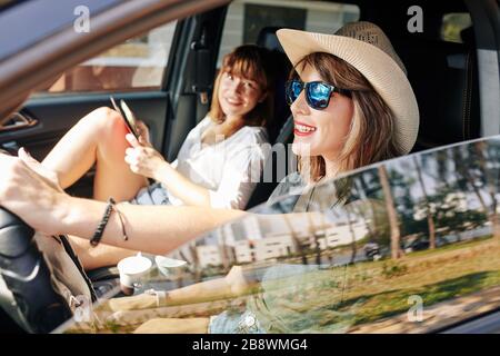 Fröhliche junge Frau in Strohhut und Sonnenbrille, die Auto fährt und mit ihrem Freund spricht Stockfoto