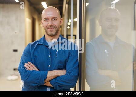 Taille hoch Porträt des reifen Glatzers lächelt an der Kamera, während er mit den Armen gestreift steht und sich selbstbewusst an die Wand lehnt, kopiere Raum Stockfoto