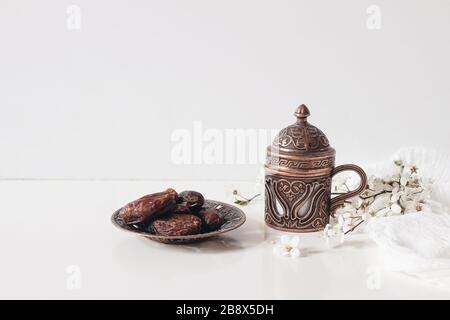 Ramadan Kareem Grußkarte, Einladung. Ziertee, Kaffeetasse, Bronzeplatte mit Datteln Obst, weißer blühender prunusbaum Zweig auf weißem Tisch. Stockfoto