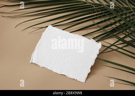 Sommerhochzeit Schreibwaren Mock-up, Schreibtisch-Szene. Leere Grußkarte, Visitenkarte und grüne Palmblätter auf beigefarbenem Tischhintergrund. Tropisch exotisches Design p Stockfoto