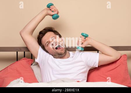 Der gutaussehende junge Mann, der vor der Hantelfront steht, hebt sich beim Training zu Hause an, während er im Bett während der Quarantäne ruht. Das Konzept "zu Hause bleiben" Stockfoto