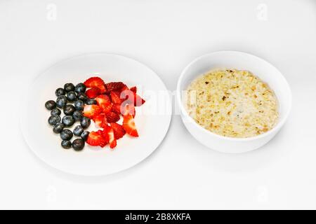 Ein gesundes, glückliches Haferflohfrühstück für eine Zeit der Isolation. Eine weiße Schüssel Hafer und ein weißer Teller frisch geschnittener Erdbeeren und mehrere Blaubeeren Stockfoto