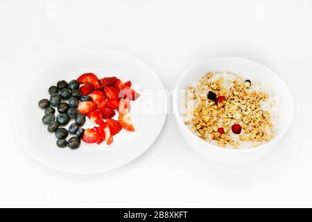 Ein gesundes, glückliches Haferflohfrühstück für eine Zeit der Isolation. Eine weiße Schüssel mit Hafer, Joghurt und Granola und eine weiße Platte mit frisch geschnittenen Erdbeeren Stockfoto