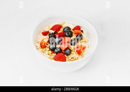 Ein gesundes, glückliches Haferflohfrühstück für eine Zeit der Isolation. Eine Schüssel Hafer, Granola, Erdbeere, Blaubeeren, Joghurt, weiße Schüssel mit weißem Hintergrund Stockfoto