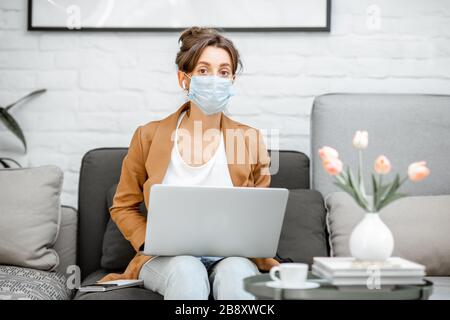 Portrait einer traurigen Frau in medizinischer Maske, die online von zu Hause aus auf dem Laptop arbeitet. Bleiben Sie während eines Virenpandemic-Konzepts zu Hause Stockfoto