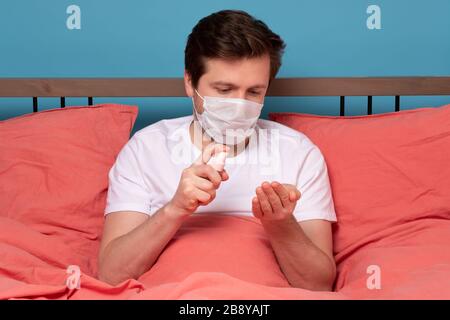 Kaukasischer Mann mit medizinischer Maske desinfiziert seine Hände mit Desinfektionsmittel aus einer Flasche, die im Bett auf Quarantäneisolierung sitzt Stockfoto