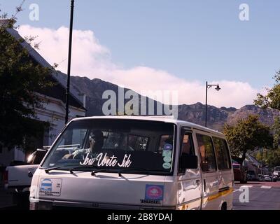 Fahrer mit leerem Taxi mit den Worten "jou voete jik" auf der Windschutzscheibe, der die Hauptstraße von Franschhoek im Westkaper, Südafrika, hinunterfährt. Stockfoto