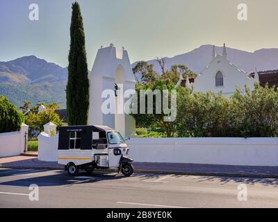 Hochauflösendes Foto eines leeren Tuk Tuk, das vor der alten reformierten holländischen Kirche aus dem Jahr 1847 in Franschhoek, Westkap, Südafrika, geparkt ist. Stockfoto