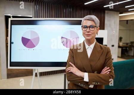 Taillen-Up-Portrait des modernen, ausgereiften Business Managers, der die Kamera betrachtet, während er während der Marketingpräsentation mit den Armen gegen das digitale Board steht Stockfoto