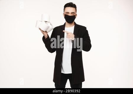 Gutaussehender junger Mann in schwarzer Schutzmaske, der vier Rollen Toilettenpapier hält und mit dem Finger darauf zeigt, isoliert auf dem weißen Hintergrund. Junge Männer Stockfoto