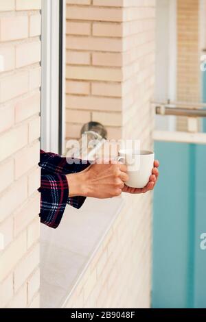 Nahaufnahme eines jungen kaukasischen Mannes, in Pyjamas, mit Kaffee am Geländer eines Fensters, in seinem Haus oder in einem Hotel Stockfoto