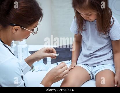 Süßes kleines Mädchen bei der Kinderärztebestellung, nette junge Frau Ärztin in einem weißen medizinischen Labormantel beschwert sich eine Bandhilfe am Knie des Mädchens Stockfoto