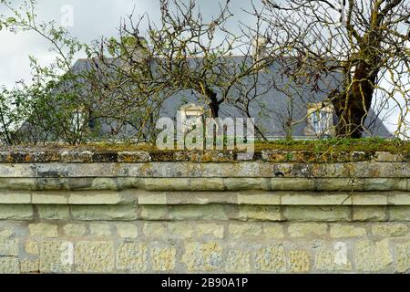 Mittelalterliche Steinmauer mit wachsendem Moos oben, im Hintergrund altes traditionelles französisches Landhaus mit Schieferdach. Stockfoto