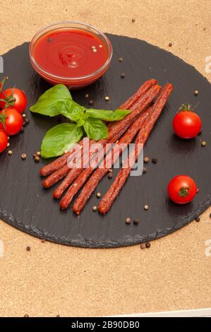 Dünne lange Wurst mit Sauce, Kirschtomaten und Basilikum auf einem schwarzen Graphitbrett Stockfoto