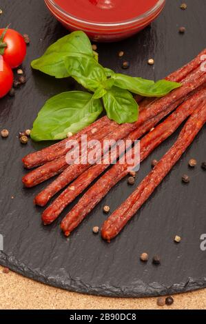 Dünne lange Wurst mit Sauce, Kirschtomaten und Basilikum auf einem schwarzen Graphitbrett Stockfoto