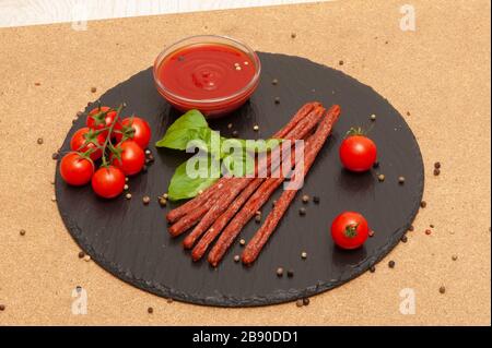 Dünne lange Wurst mit Sauce, Kirschtomaten und Basilikum auf einem schwarzen Graphitbrett Stockfoto