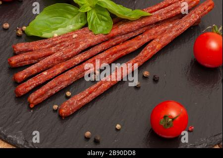 Dünne lange Wurst mit Sauce, Kirschtomaten und Basilikum auf einem schwarzen Graphitbrett Stockfoto