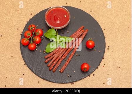 Dünne lange Wurst mit Sauce, Kirschtomaten und Basilikum auf einem schwarzen Graphitbrett Stockfoto