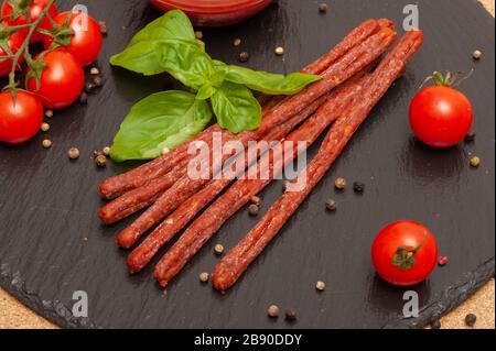 Dünne lange Wurst mit Sauce, Kirschtomaten und Basilikum auf einem schwarzen Graphitbrett Stockfoto