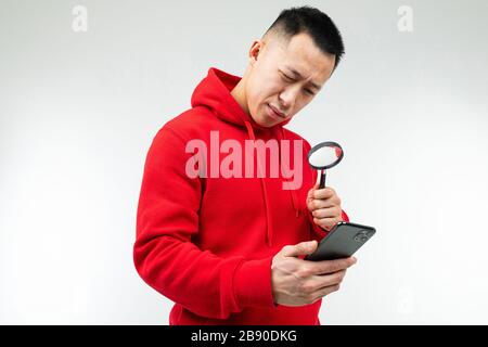 Brünette Kerl in einem roten Pullover schaut durch eine Lupe auf weißem Hintergrund auf das Telefon Stockfoto