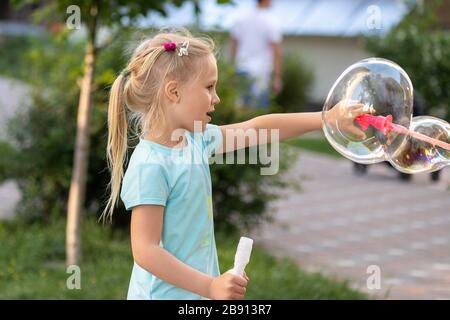Portrait des kleinen süßen kaukasischen blonden Mädchens, das Spaß und Freude hat, große Seifenblasen zu blasen, die auf dem städtischen Straßenpark draußen spielen. Fröhliches liebensliches Kind Stockfoto