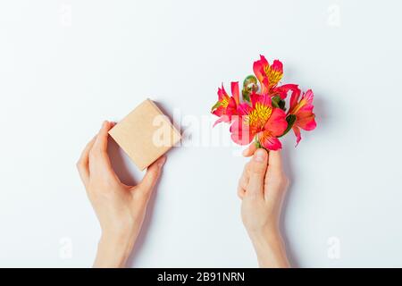 Von oben Frauenhände mit kleiner Geschenkbox und Blumenstrauß mit frischen Blumen auf weißem Hintergrund, flacher Lay. Stockfoto
