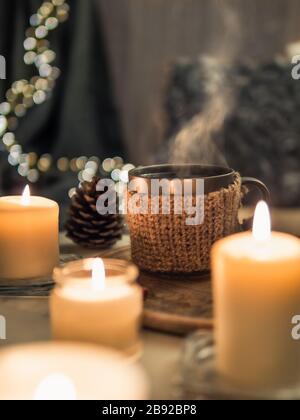 Gemütliches Zuhause, Hyge, Gemütlichkeit - heißes Getränk in der Tasse mit Dampf und brennenden weißen Duftkerzen in der Nähe des Sofas mit Kissen und Plaid. Winterdekor und heiße Teetasse. Vertikal Stockfoto