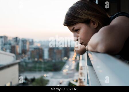 Junge traurige Frau, die durch den Balkon eines Apartmentgebäudes nach draußen blickt Stockfoto