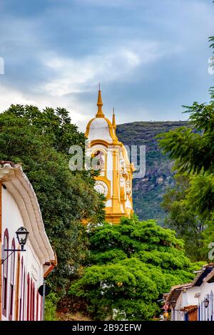 Historischer Kirchturm aus dem 18. Jahrhundert, der durch Bäume und Häuser in der Altstadt von Tiradentes in Minas Gerais, Brasilien, gesehen wird Stockfoto