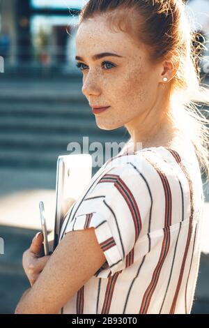 Nahaufnahme des Porträts einer kaukasischen Frau mit Freckeln und roten Haaren, die ihren Laptop bei einem Spaziergang halten Stockfoto