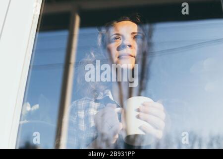 Junge blonde Frau, die mit einem besorgten Gesichtsausdruck aus dem Fenster schaut. Spiegelung im Glas, doppelter Belichtungseffekt Stockfoto