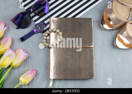 Ein goldenes Notizbuch mit Damenschuhen, Tulpen, Brille und einer Uhr auf konkretem Hintergrund. Fleet lag Stockfoto