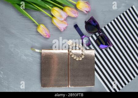 Ein goldenes Notizbuch mit Damenschuhen, Tulpen, Brille und einer Uhr auf konkretem Hintergrund. Fleet lag Stockfoto