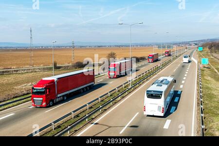 Der Konvoi der roten Transportfahrzeuge führt zwei weiße Busse auf einer Landstraße auf dem Land unter einem wunderschönen blauen Himmel vorbei Stockfoto