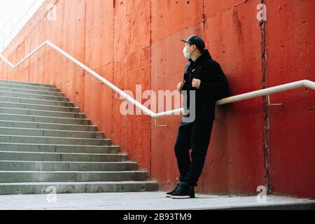 Ein junger Mann in schwarzer Sportswear und eine Baseballkappe mit einer medizinischen Maske auf seinem Gesicht nahe der roten Wand. Schutz vor dem Virus Stockfoto