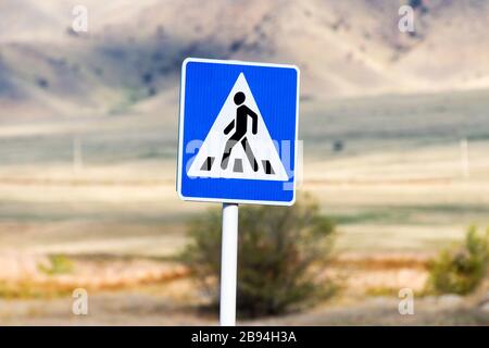 Weißes und blaues Straßenschild, das eine Person zeigt, die in Sicherheit den Fußgängerüberweg überquert. Straßenschild mit Hintergrund der Berge aus Fokus in Kirgisistan, Stockfoto